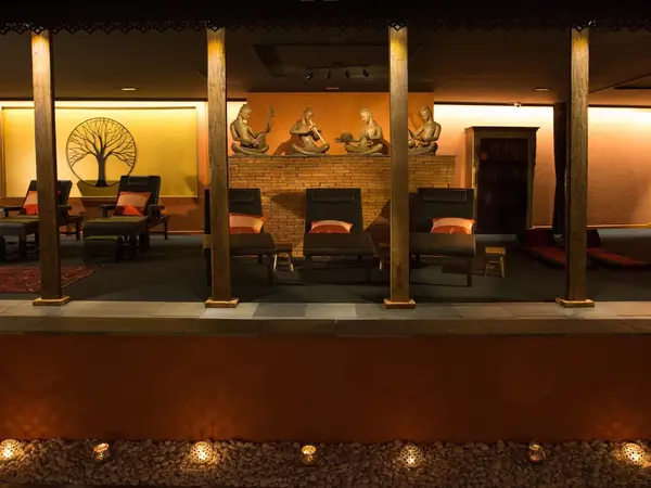 Warm spa lounge with loungers, wood pillars, statues, and soft ambient lighting
