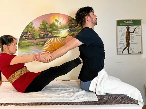 Two people perform Thai massage stretches on a treatment bed, with wall art and anatomy poster
