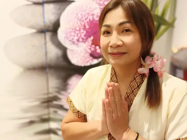 Smiling woman in Thai massage attire, hands together, with floral wall decor