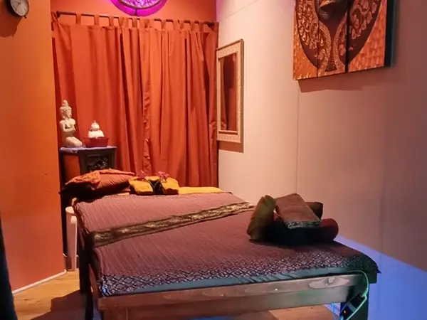 Cozy massage room with a treatment bed, orange curtains, and Thai decor