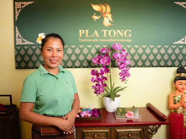 Smiling woman stands in a Thai massage salon with orchids and decorative statue