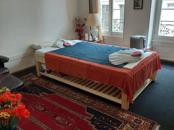 A serene massage room with a treatment bed, rugs, lamp, and windows