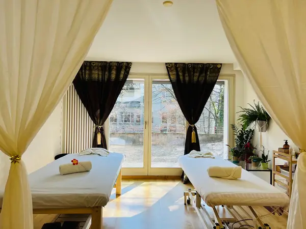 Bright massage room with two tables, curtains, plants, and sunlight through windows