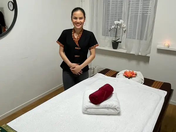 Bright massage room with a smiling therapist beside a neatly made treatment bed