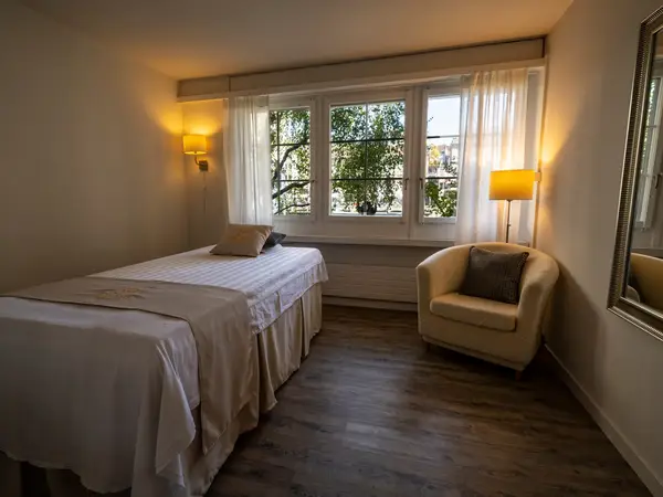 Cozy treatment room with massage bed, armchair, lamps, and window light