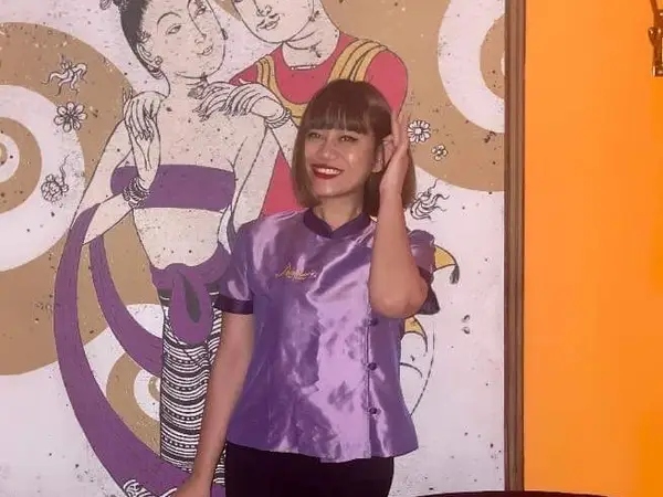 Woman in purple uniform stands before Thai-themed mural and orange wall