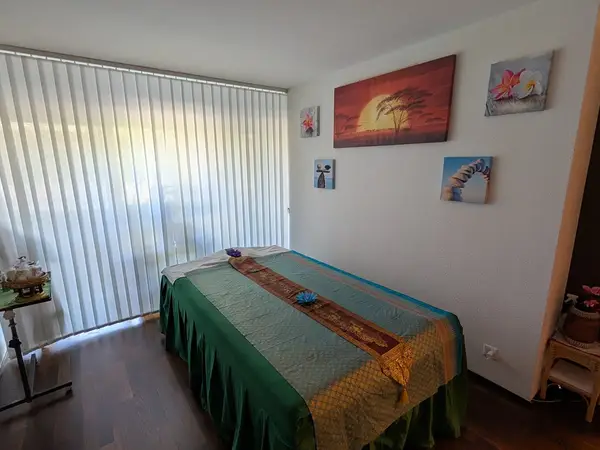 Bright massage room with bed, vertical blinds, and tropical wall art