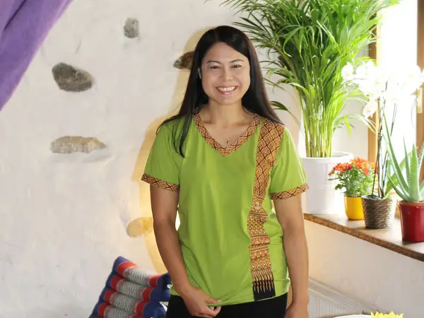 Smiling woman stands in a bright Thai massage room with potted plants and cushions