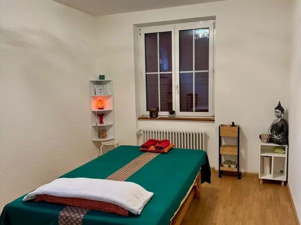 Massage room with green-covered table, white walls, window, and Buddha decor