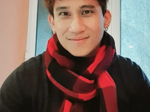 A smiling person indoors wearing a red-and-black scarf near a window