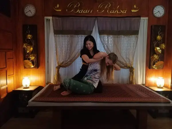 Warm Thai massage room with two women on a raised treatment bed, lit by lamps