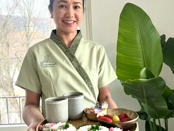 Smiling woman in Thai massage uniform holding a tray beside a plant by the window